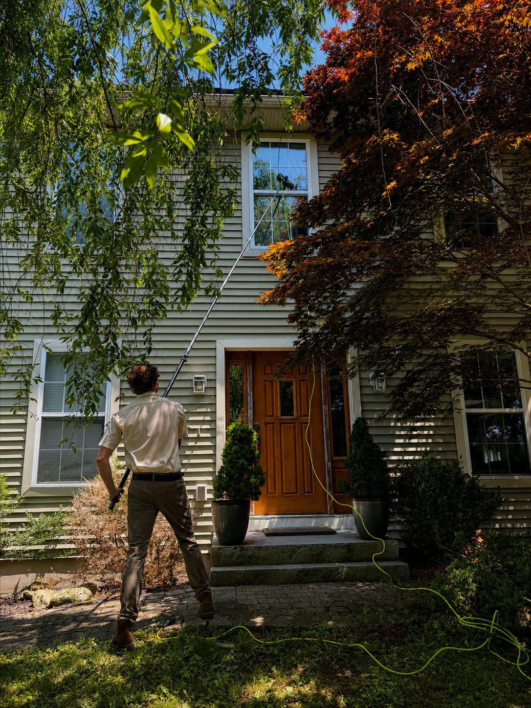 Man cleaning a second-story window with a long extension pole on a suburban house.