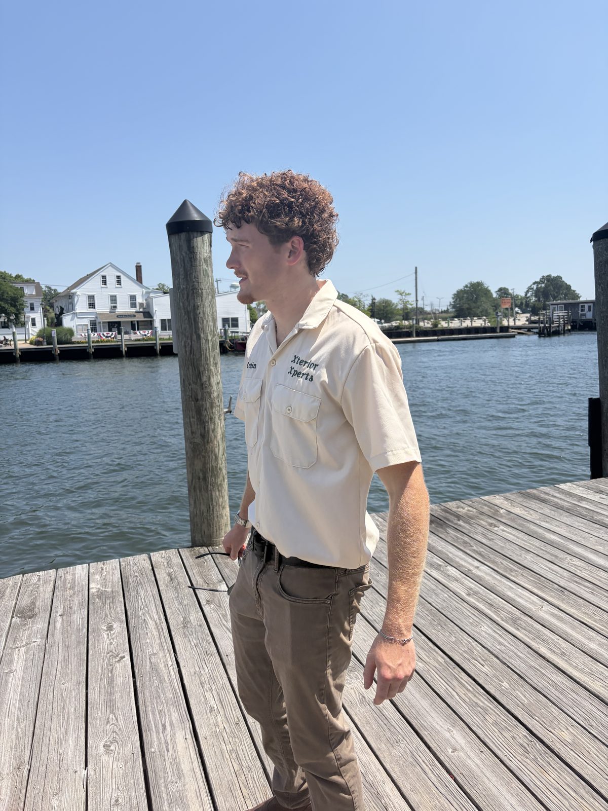 Man with curly hair in a work shirt stands on a wooden dock by water.